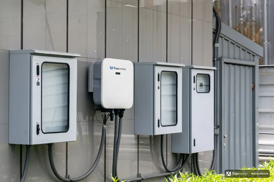 Białe urządzenie na elewacji budynku między szarymi szafkami elektrycznymi, pod nim przewody w osłonach. Na froncie naniesiono logo TraxSystem i okienko z trzema diodami statusu.