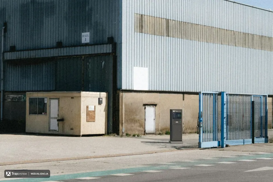 Wjazd na betonowy plac przed halą magazynową w słoneczny dzień: kontener portierni, niebieska brama i ulica z pasem rowerowym. Naniesiono kiosk kierowcy z logo TraxSystem i ekranem kolejki przy bramie oraz małą skrzynkę urządzenia brzegowego ANPR na bocznej ścianie portierni.
