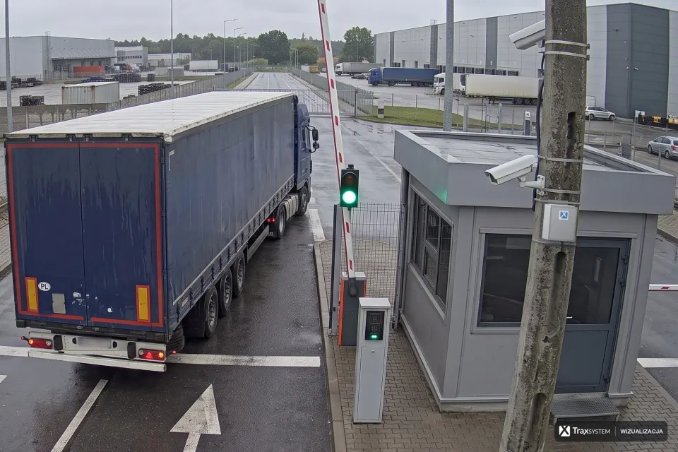 Ciężarówka z naczepą wyjeżdża pod podniesionym szlabanem obok budki ochrony. Na betonowym słupie pod dwiema kamerami naniesiono szarą skrzynkę urządzenia brzegowego z naklejką z sygnetem TraxSystem, zamocowaną dwiema metalowymi opaskami; tablica rejestracyjna naczepy jest rozmyta.
